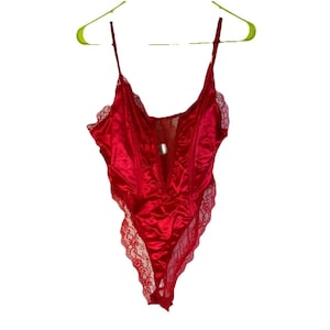 May include: A red satin bodysuit with delicate lace trim along the edges. The lingerie features adjustable straps and a deep V-neckline. The garment is hanging on a green hanger against a white background.