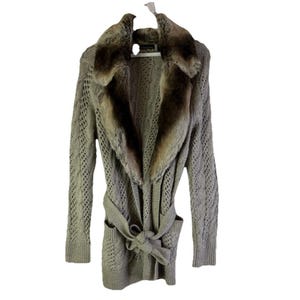 Könnte beinhalten: Ein hellgrauer Strickcardigan mit einem Kunstfellkragen und einem passenden Bindegürtel. Der langärmlige Pullover hat ein strukturiertes Open-Knit-Design und zwei Vordertaschen. Der Cardigan hängt an einem weißen Kleiderbügel.