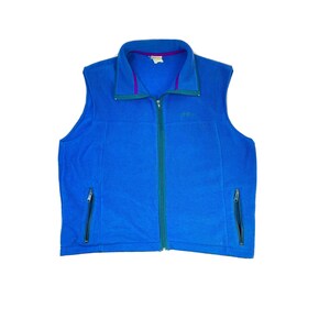 Peut inclure: Un gilet en polaire bleu avec une fermeture éclair et deux poches zippées. Le gilet a une fermeture éclair verte et une doublure violette autour du col.