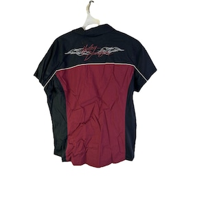 Pode incluir: Camisa preta e bordô de manga curta com acabamento branco e o logotipo da Harley-Davidson bordado nas costas. A camisa tem gola e está pendurada em um cabide branco.