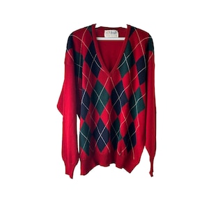 Può includere: Maglione rosso con un motivo a rombi nero, verde e oro. Il maglione ha un collo a V e maniche lunghe.