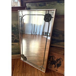 May include: A rectangular mirror with a gold-colored frame and decorative black metal accents. The mirror features a floral design at the top and bottom corners. The mirror reflects the room and is set against a backdrop of artwork.