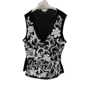 Op de afbeelding: Zwart mouwloos topje met een V-hals en ingewikkelde zilveren pailletten bloemen- en krulversieringen. De top is gemaakt van een doorschijnend zwart mesh materiaal en hangt aan een witte hanger.