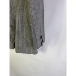 Puede incluir: Un blazer gris con un sutil estampado de rayas. El blazer tiene tres botones en el pu&ntilde;o de la manga. La tela parece ser un material tejido. El blazer est&aacute; colgado sobre un fondo blanco.