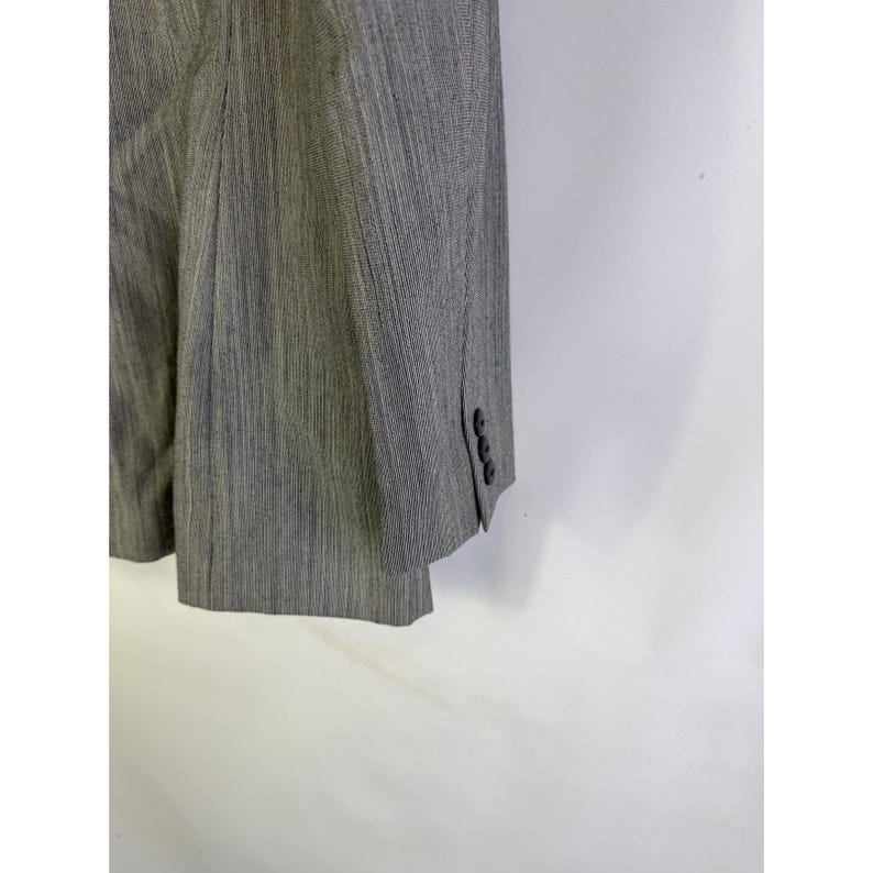 Puede incluir: Un blazer gris con un sutil estampado de rayas. El blazer tiene tres botones en el pu&ntilde;o de la manga. La tela parece ser un material tejido. El blazer est&aacute; colgado sobre un fondo blanco.