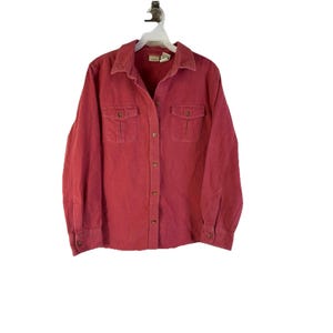 Puede incluir: Una camisa roja de manga larga con dos bolsillos en el pecho y botones. La camisa está colgada de una percha blanca sobre un fondo blanco. Tiene cuello y puños abotonados.