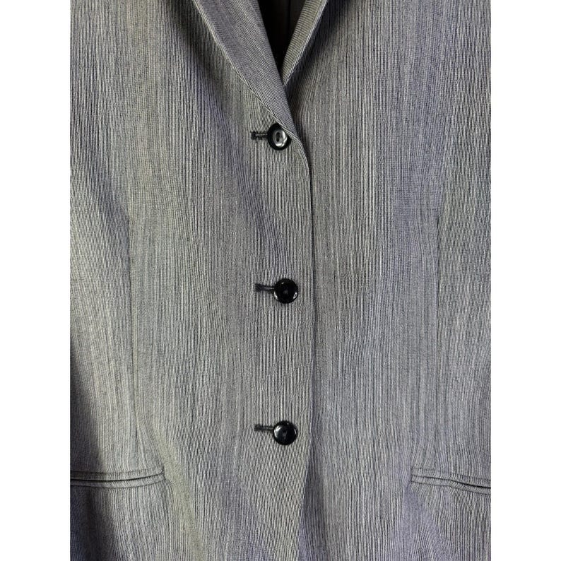 Puede incluir: Primer plano de un blazer gris con tres botones negros. El blazer tiene una tela texturizada y solapas con muescas. Los botones son redondos y brillantes, contrastando con la tela mate.