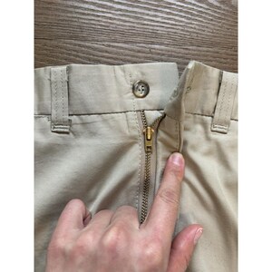 May include: Close-up of the zip and waistband of a pair of beige trousers. The zip is gold metal and the waistband has a single button.