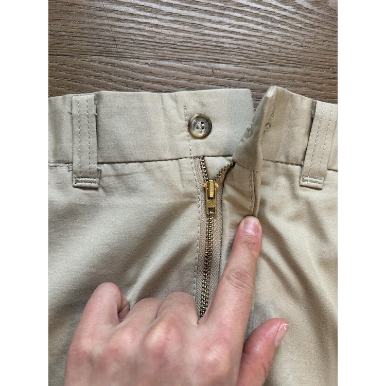 May include: Close-up of the zip and waistband of a pair of beige trousers. The zip is gold metal and the waistband has a single button.