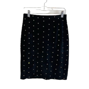 Puede incluir: Una falda de terciopelo negro con adornos dorados. La falda es de largo hasta la rodilla y tiene una silueta ajustada.