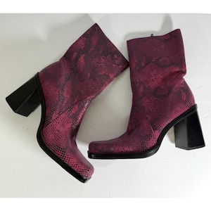 May include: A pair of ankle boots featuring a magenta and black snakeskin pattern. The boots have a square toe design, a mid-calf height, and a thick, black block heel. The construction includes a side zipper closure on the inner side of each boot.