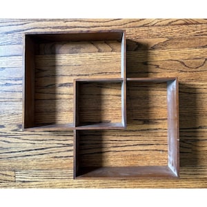 May include: A wall-mounted wooden shelf unit featuring three interconnected square and rectangular open compartments. The structure is crafted from medium-toned brown wood with a visible grain, creating a geometric arrangement of open storage spaces.