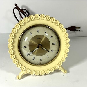 May include: Cream-coloured vintage clock with a floral design around the face. The clock has a gold-toned face with black Roman numerals and hands. The clock has a brown power cord and plug.