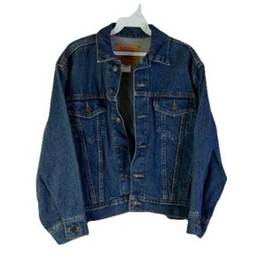 May include: A classic blue denim jacket with a button-up front, two chest pockets, and side pockets. The jacket is hanging on a white hanger. The jacket has a collar and buttoned cuffs. The denim is a medium wash.