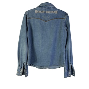 Może przedstawiać: Jasnoniebieska koszula dżinsowa z długimi rękawami i mankietami zapinanymi na guziki. Tył koszuli zdobi haftowany napis "HARLEY-DAVIDSON" nad dekoracyjnym wzorem. Koszula ma klasyczny kołnierzyk i karczek w stylu western.