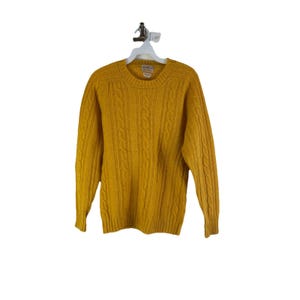 May include: Mustard yellow cable knit sweater with a crew neck. The sweater is hanging on a white hanger against a white background. The sweater has a textured cable knit design and long sleeves.