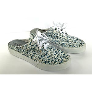 Op de afbeelding: Een paar canvas sneakers met bloemenprint, witte veters en een witte rubberen zool. De schoenen hebben een crèmekleurige achtergrond met een patroon van kleine blauwe bloemen.
