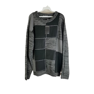 May include: A long-sleeved sweater with a patchwork design. The sweater features a crew neck and a mix of gray and black panels. A tag hangs from the sweater. The sweater is hanging on a white hanger.