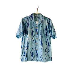 May include: Light blue short-sleeve shirt with a pattern of dark blue shark-like figures. The shirt has a collared neckline, a front pocket, and wooden buttons. The shirt is hanging on a gold-colored hanger.