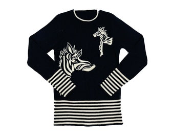 Può includere: Un maglione nero a maniche lunghe con polsini e fascia inferiore a righe bianche. Il maglione presenta una grafica bianca di due zebre sul davanti.