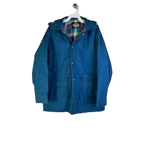 Puede incluir: Una chaqueta azul vintage con capucha y múltiples bolsillos. La chaqueta tiene un forro de cuadros y una cintura con cordón. La chaqueta está colgada de una percha blanca.