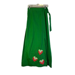 Può includere: Una gonna a portafoglio verde brillante con chiusura a fiocco. La gonna presenta tre applicazioni a forma di cuore rosso con dettagli a scacchi verdi e bianchi e cuciture nere. Appesa a una gruccia bianca.