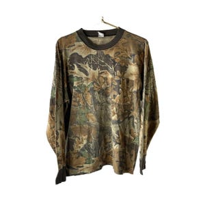 Könnte beinhalten: Langärmeliges Camouflage-Shirt mit Rundhalsausschnitt und Brusttasche. Das Shirt hat ein braunes, grünes und beiges Blättermuster. Die Manschetten und der Kragen sind dunkelbraun. Das Shirt hängt an einem Metallbügel.