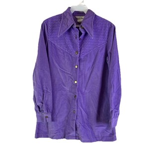 May include: A vintage purple corduroy shirt with a pointed collar and long sleeves. The yoke features a quilted design, and the shirt fastens with gold-coloured buttons. The cuffs also have button closures. The shirt is hanging on a white hanger.