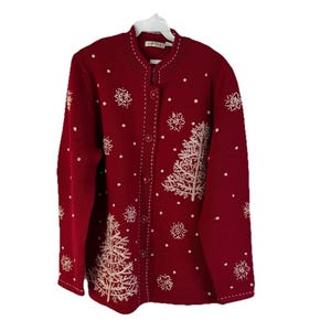 Könnte beinhalten: Ein roter Cardigan mit weißen, gestickten Schneeflocken und Weihnachtsbäumen. Der Pullover hat eine Knopfleiste vorne und einen runden Ausschnitt. Der Markenname "Orvis" ist auf dem Etikett zu sehen.