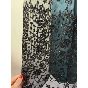 May include: Close-up of a black lace fabric with a floral pattern. The lace is layered over a teal fabric, creating a contrast in color and texture. The intricate design of the lace is visible, with delicate details and a shimmering effect.