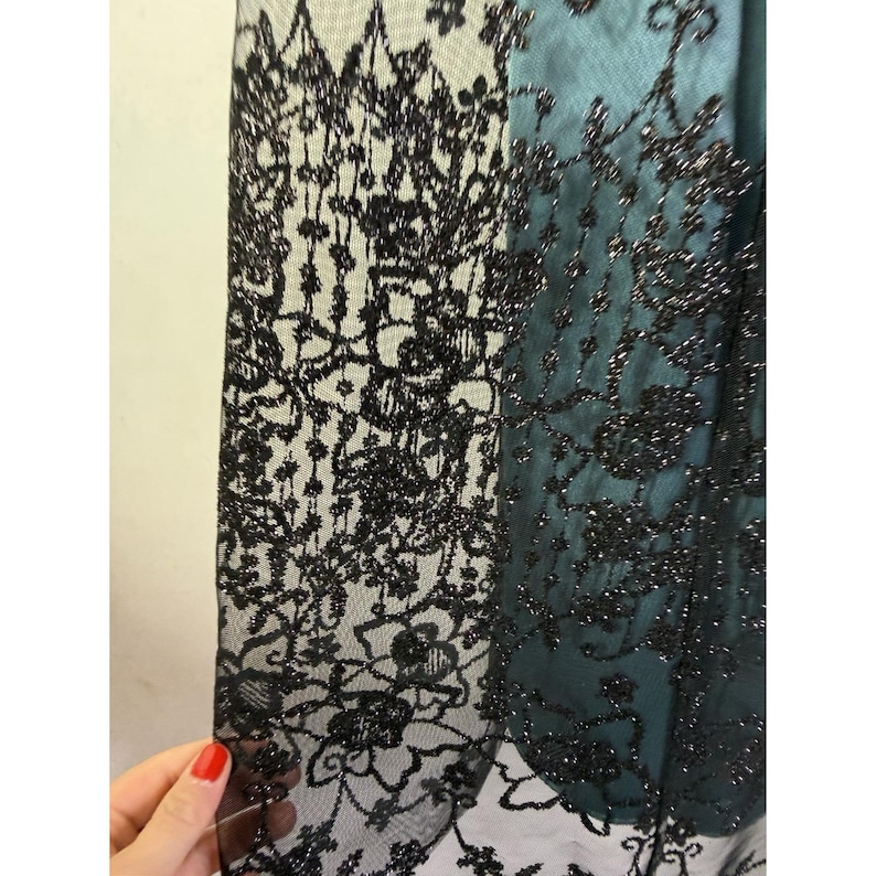 May include: Close-up of a black lace fabric with a floral pattern. The lace is layered over a teal fabric, creating a contrast in color and texture. The intricate design of the lace is visible, with delicate details and a shimmering effect.