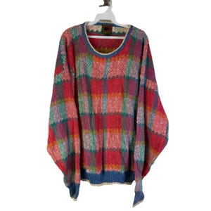 Könnte beinhalten: Ein übergroßer Pullover mit einem farbenfrohen Karomuster in Rot-, Rosa-, Grün- und Blautönen. Der Pullover hat einen runden Halsausschnitt mit blauem Besatz und lange Ärmel. Er hängt an einem weißen Kleiderbügel.