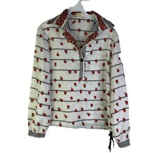 May include: White long-sleeve blouse with a floral pattern. The blouse features red roses and black zigzag stripes. The collar and cuffs have a black and white zigzag pattern. A single black button is at the neckline.