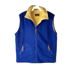 Puede incluir: Un chaleco de forro polar azul con forro de polar amarillo. El chaleco tiene cierre de cremallera y dos bolsillos con cremallera. El chaleco tiene una etiqueta "Pendleton" marrón en la parte delantera.