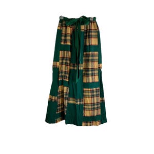 May include: A long, patchwork skirt featuring a combination of green and plaid fabric. The skirt has a high waist with a green tie. The plaid pattern includes shades of tan, red, and black. The skirt is designed to be worn at the waist.