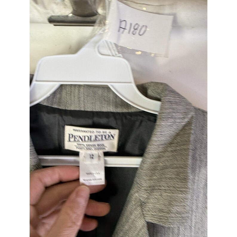 Puede incluir: Un blazer gris Pendleton, talla 12, cuelga de una percha blanca. El blazer tiene un aspecto texturizado y un forro oscuro. La etiqueta Pendleton es visible, junto con una etiqueta con el n&uacute;mero A180.