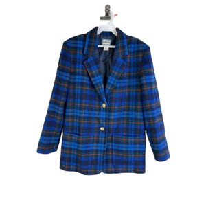 Puede incluir: Una chaqueta azul de cuadros con botones dorados. La chaqueta tiene dos bolsillos en la parte delantera.