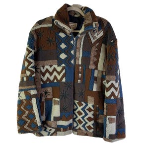 May include: A patchwork fleece jacket in shades of brown, blue, and cream. The jacket features a high collar, a full-length zipper, and a variety of geometric and abstract patterns. The jacket is hanging on a white hanger.