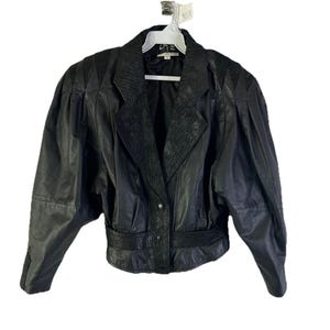 May include: Black leather jacket featuring a notched collar, shoulder pads, and a belted waist. The jacket has a textured panel on the collar and a button closure. The sleeves are gathered at the shoulders.