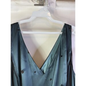May include: A teal-colored formal dress with a V-neck design, hanging on a white hanger. The dress features a sheer black overlay with scattered dark embellishments. The dress is sleeveless and appears to be made of a silky material.