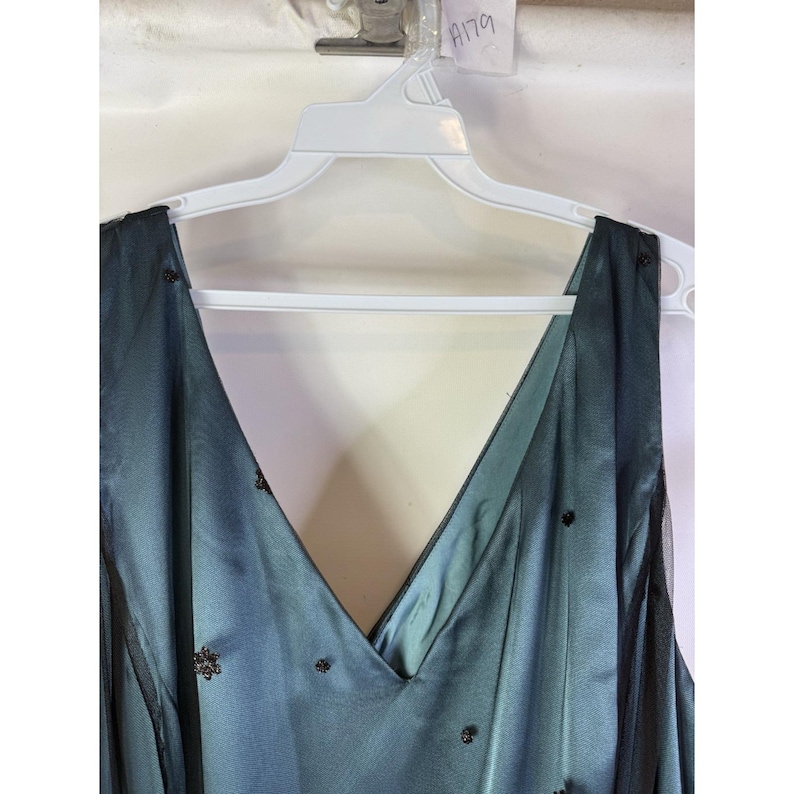 May include: A teal-colored formal dress with a V-neck design, hanging on a white hanger. The dress features a sheer black overlay with scattered dark embellishments. The dress is sleeveless and appears to be made of a silky material.