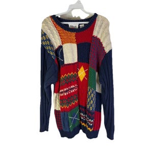 May include: A patchwork sweater with a variety of colors and patterns. The sweater features a mix of red, blue, yellow, and cream-colored blocks. The sleeves are a solid navy blue. The sweater is hanging on a white hanger.