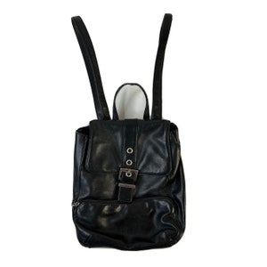 Puede incluir: Una mochila de cuero negro con una hebilla plateada y dos correas ajustables.