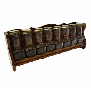 Peut inclure: Un porte-épices en bois avec huit bocaux en verre remplis d'épices. Les bocaux sont étiquetés avec les noms des épices : Sel, Sel de mer, Sauge, Paprika, Citron, Moutarde, Crm. Tar et Cannelle.