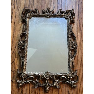 May include: Ornate rectangular mirror with a bronze-colored frame. The frame features detailed floral and scrollwork designs. The mirror is set against a wooden background.
