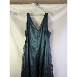 May include: A teal-colored formal dress with a V-neck and a flowing silhouette. The dress features a sheer overlay with black floral embroidery along the hemline. It hangs on a white hanger against a white background.