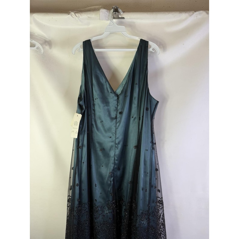 May include: A teal-colored formal dress with a V-neck and a flowing silhouette. The dress features a sheer overlay with black floral embroidery along the hemline. It hangs on a white hanger against a white background.