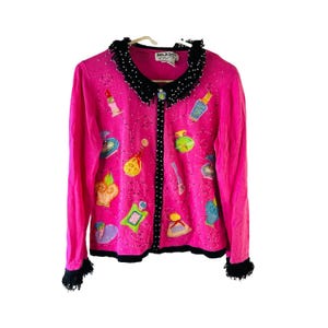 May include: A pink cardigan with a black trim and a black button closure. The cardigan is decorated with colorful embroidered images of perfume bottles and lipstick.