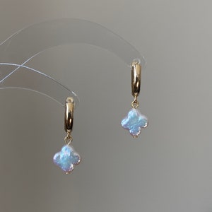 Pode incluir: Um par de brincos de argola dourados, cada um com um pequeno pingente iridescente em forma de trevo de quatro folhas. Os pingentes têm um acabamento perolado branco-azulado e estão suspensos em argolas de metal dourado polido.