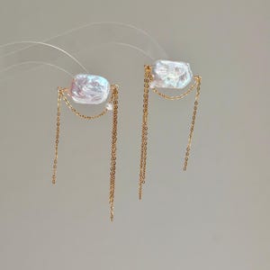 May include: A pair of gold-toned earrings featuring rectangular, iridescent white baroque pearls. Each earring is accented with delicate gold chains that drape beneath the pearl, creating a layered, dangling effect with a polished finish.